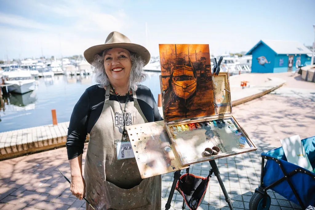An artist stands beside an easel displaying a painting of a boat, with Mandurah's waterfront and marina in the background.